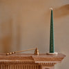 A tall dark green taper advent candle with numbered days from 1 to 24 printed vertically, displayed on a small round wooden base on a wooden mantelpiece. A brass candle holder is visible in the background against a warm beige wall.