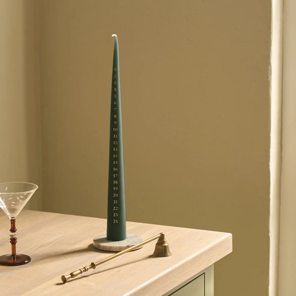 Green tapered advent candle sits on marble candle plate next to a brass candle snuffer, on a wooden counter.