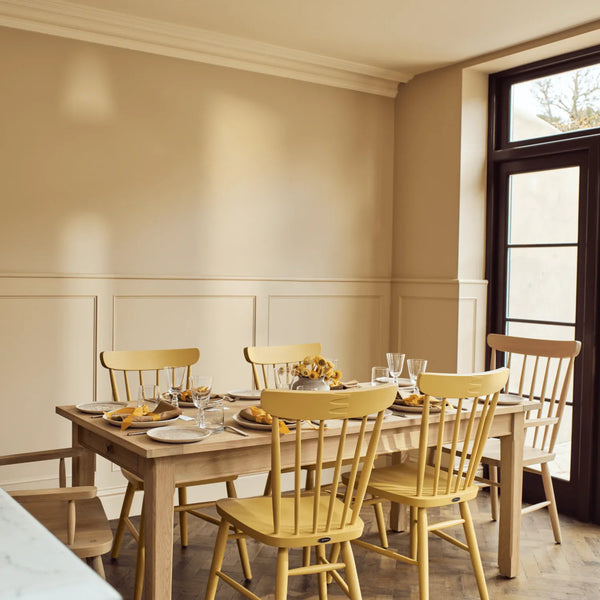A dining room scene with a light wood rectangular table surrounded by six yellow spindle-back chairs, set with plates and glasses near a large dark-framed window.