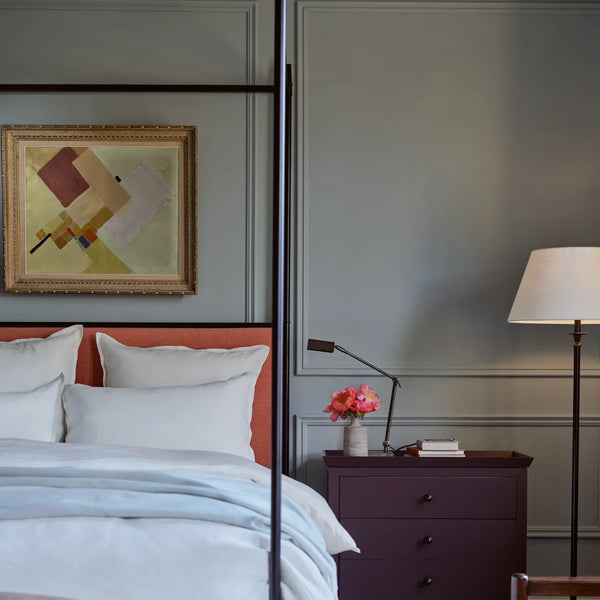 A styled bedroom interior featuring a bed with white bedding and a rust-orange headboard, set against a gray paneled wall with a framed abstract painting. A dark purple nightstand, a black floor lamp with a white shade, and a desk lamp with a pink flower arrangement are visible.