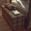 Eloise ottoman upholstered in Harris Tweed Fig, styled in a warm interior with a folded blanket and books resting on its checked upholstered surface.