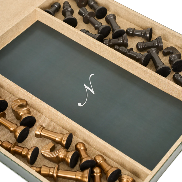 A close-up of a monogrammed chess set in a presentation box, featuring a dark green central plaque with a white cursive monogram 'N' and bronze and black chess pieces arranged in compartments.