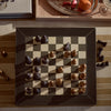 A square leather chess board with a dark brown border and cream-and-dark checkered center, holding a set of chess pieces in black and bronze tones. The board is photographed from above on a striped beige fabric surface with a few chess pieces placed around it.