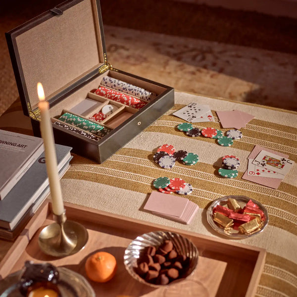 An open luxury card and chip box displayed on a striped tablecloth with playing cards, poker chips, candy, and a lit candle in a warm, elegant setting.