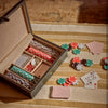 An open luxury poker set storage box with multiple compartments holding stacks of colorful poker chips, shown on a striped beige tablecloth with scattered chips and playing cards nearby.