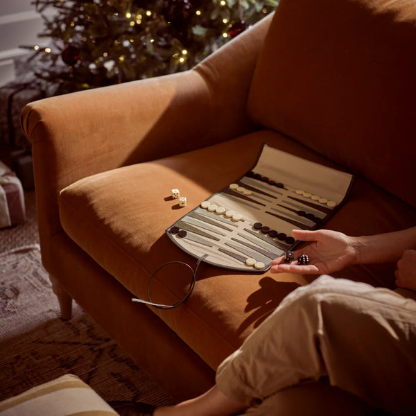 A backgammon set laid open on an orange-brown armchair in a warmly lit living room, with a person's hand holding dice nearby and a blurred Christmas tree in the background.