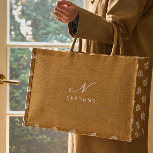A person in a tan coat holds a large mustard yellow jute-style shopping bag with white floral trim and a white NEPTUNE logo on the front, photographed indoors near a window.