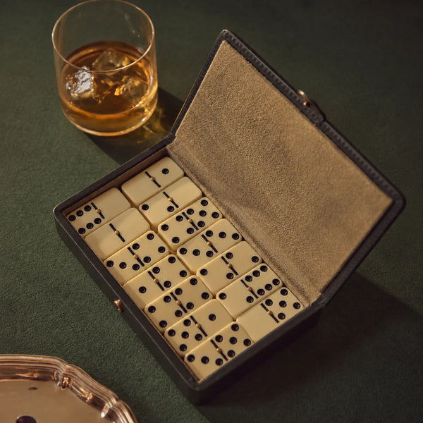An open black leather domino game box with cream-colored tiles, styled on a dark green table next to a glass of whiskey.