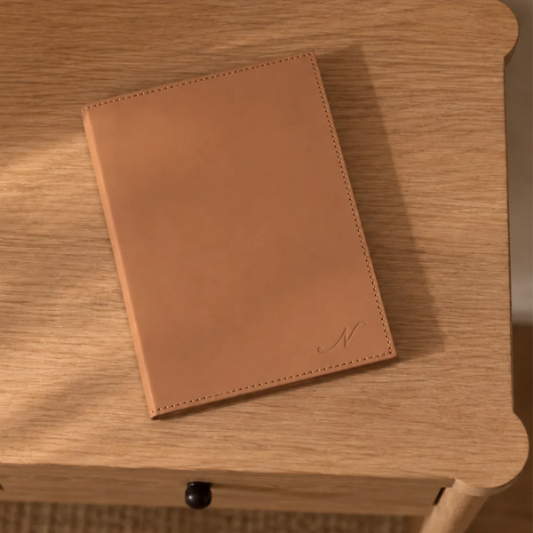 A tan leather notebook with stitched edges and a small embossed monogram, resting on a light wood table.