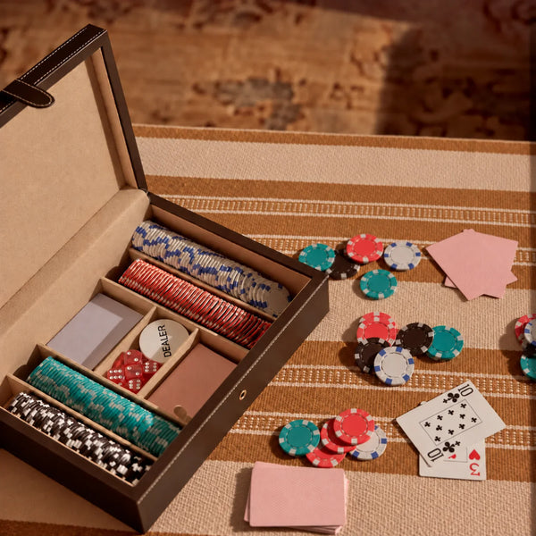 An open luxury poker set storage box with multiple compartments holding stacks of colorful poker chips, shown on a striped beige tablecloth with scattered chips and playing cards nearby.