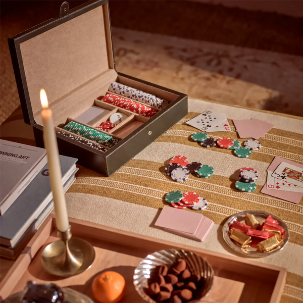 An open luxury poker set storage box with multiple compartments holding stacks of colorful poker chips, shown on a striped beige tablecloth with scattered chips and playing cards nearby.