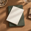 A white notebook refill resting on top of a dark green leather journal with stitched edges, photographed from above on a warm wood tabletop with soft natural shadows.