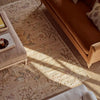 A styled interior scene showing a large rectangular area rug with an ornate distressed pattern in warm beige, tan, and muted brown tones. The rug is placed under a tan sofa and a light upholstered ottoman, with natural sunlight casting shadows across the floor.