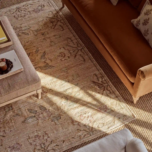 A styled interior scene showing a large rectangular area rug with an ornate distressed pattern in warm beige, tan, and muted brown tones. The rug is placed under a tan sofa and a light upholstered ottoman, with natural sunlight casting shadows across the floor.