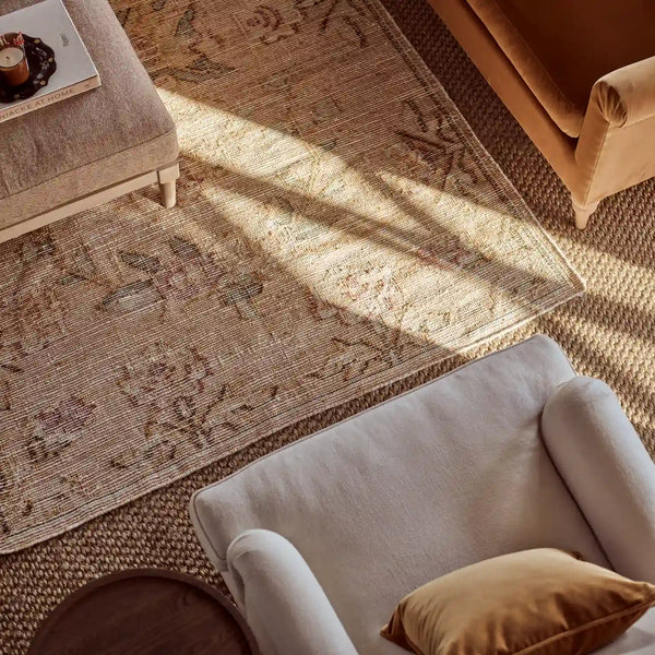 A styled interior scene showing a large rectangular area rug with a distressed floral pattern in beige, tan, and muted mauve tones. The rug is placed on a woven floor between upholstered seating pieces, with warm sunlight casting shadows across it.