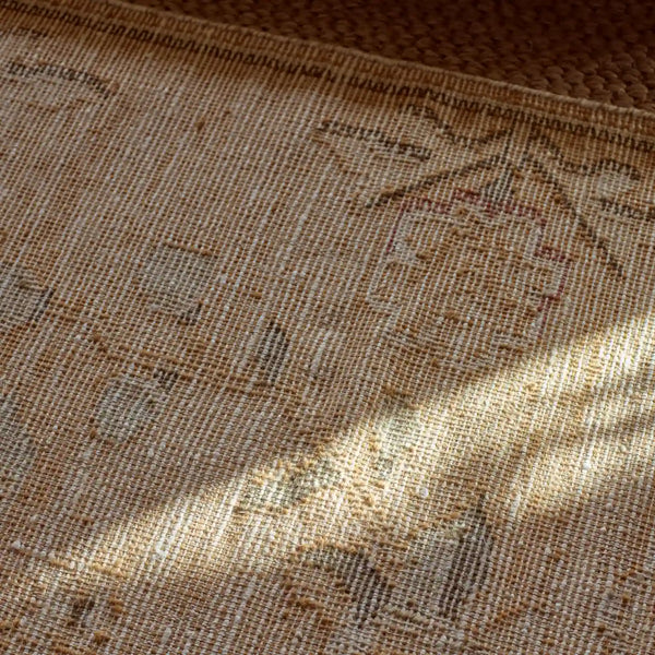 A close-up of a woven rug or textile in warm beige and tan tones with subtle faded geometric and floral motifs, lit by dappled sunlight and shadow.