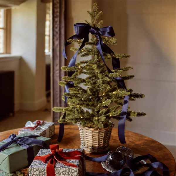 A small potted Christmas tree with gold glittery branches, decorated with a large navy blue ribbon and bow, set on a wooden table with wrapped gifts in a warm, cozy room.