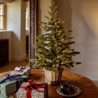 A small artificial Christmas tree in a woven basket pot, lit with warm white string lights, set on a wooden table next to wrapped gift boxes in a cozy indoor room.