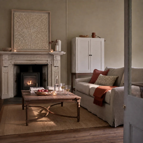 Overton wall art in a cozy living room scene with a cream slipcovered sofa, red and beige pillows, a wooden coffee table, a fireplace with a lit fire, a white cabinet, and a large framed wall art piece on a beige wall.