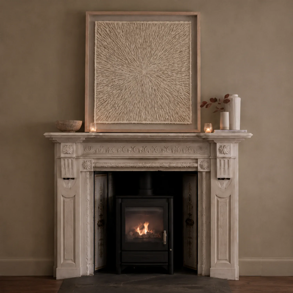Large square framed artwork with a textured radial pattern in a styled interior scene showing a carved stone fireplace mantel with a black wood-burning stove lit with a fire, set against a warm beige wall.