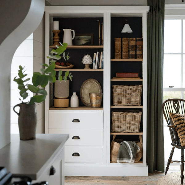 A tall white built-in shelving unit with open shelves and drawers, styled with books, wicker baskets, ceramic vessels, and decorative objects in a bright kitchen or home interior.