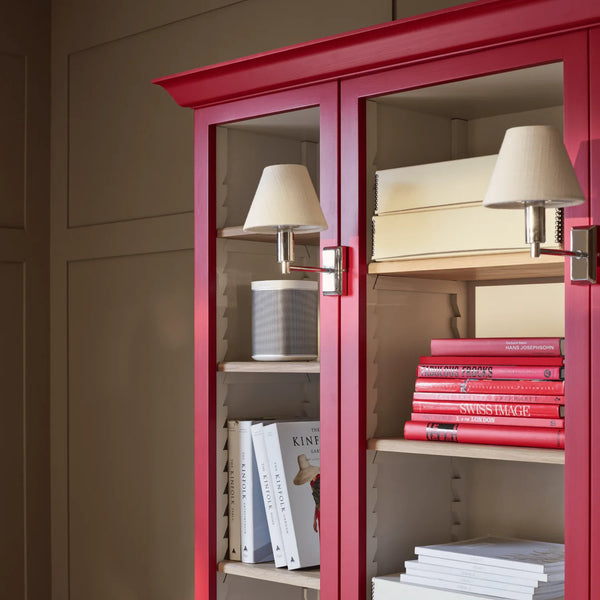 A tall red glass-front bookcase or cabinet with interior shelves, styled with books, two wall-mounted table lamps, and a small cylindrical decor object, shown against a beige paneled wall.