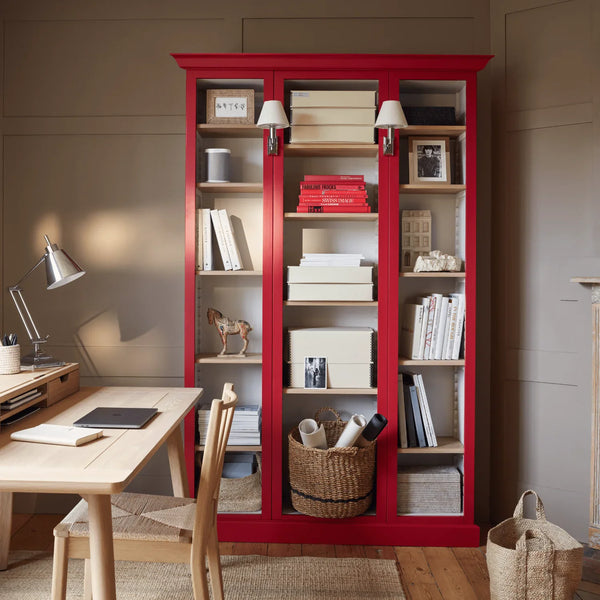 A tall red three-door bookcase with glass panels stands in a styled home office or study, holding books, boxes, and decor. A light wood desk, chair, lamp, and woven basket are visible in the room.