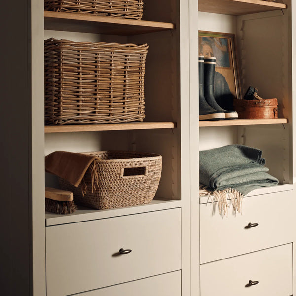 A styled closet organizer with off-white drawers and open shelving holding natural wicker baskets, folded blankets, and a pair of dark rain boots.