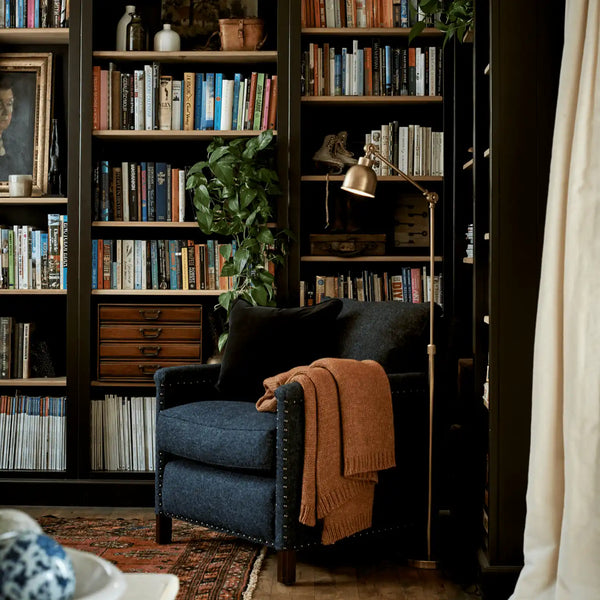 A cozy navy blue armchair in a rustic library setting, styled with a rust-orange knit throw blanket and a black pillow, positioned in front of tall dark wood bookshelves.
