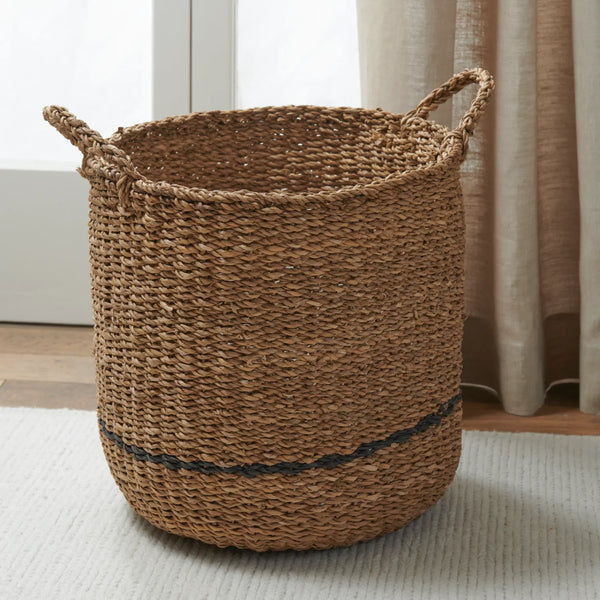 A natural woven seagrass storage basket with two rope handles and a dark horizontal stripe near the base, placed on a light rug in a softly lit room with curtains and a window behind it.