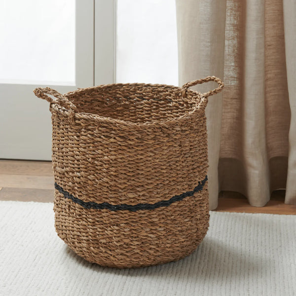 A natural woven seagrass storage basket with two rope handles and a dark navy stripe around the middle, placed on a white rug in a bright room with curtains and a window.