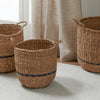 Three natural woven seagrass storage baskets with dark blue stripe accents, displayed on a light rug in a bright room with curtains.