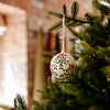 A white Christmas ornament decorated with a red and green mistletoe pattern, hanging from a green Christmas tree branch.
