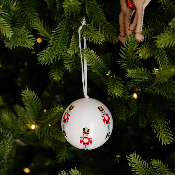 A white spherical Christmas ornament decorated with small soldier figurines, hanging from a green Christmas tree branch with warm string lights in the background.
