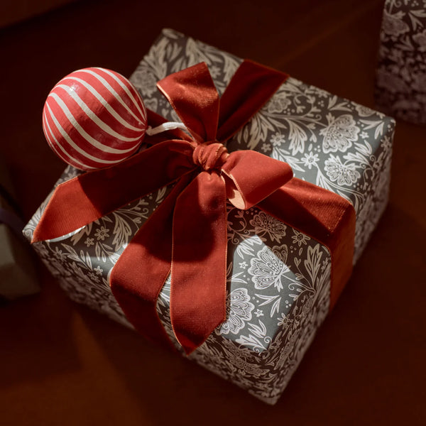 A wrapped gift box with gray and white floral-patterned paper, tied with a large rust-red satin ribbon and bow, and decorated with a red-and-white striped ornament, set on a dark wooden surface.