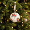 A white Christmas ornament featuring colorful sleighs carrying gift boxes, hanging from a green Christmas tree with warm string lights.