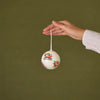 A hand holding a small white Christmas ornament with colorful sleigh illustrations against a solid olive green background.