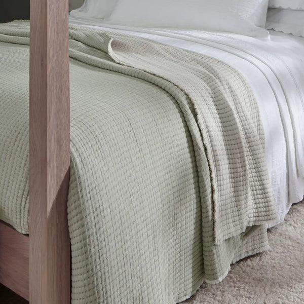 A pale sage green textured waffle-weave throw blanket draped over a bed with white bedding and a wooden bed frame.