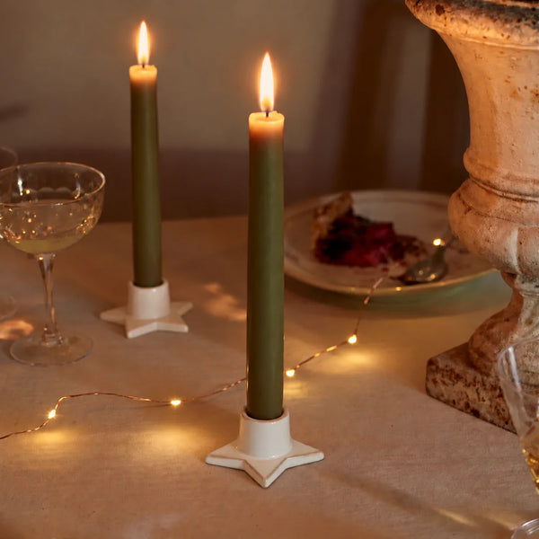 Two lit olive green taper candles in white star-shaped holders on a table, styled with warm fairy lights, a coupe glass, and a plate in a softly lit indoor setting.