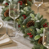 A festive holiday table garland made of green pine branches, brown pinecones, red berries, and small warm white string lights, styled on a beige tablecloth with clear drinking glasses and a folded cream napkin.