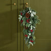 A hanging Christmas garland with green pine branches, red berries, white eucalyptus leaves, and brown pinecones, suspended from a gold hook on an olive green paneled door.
