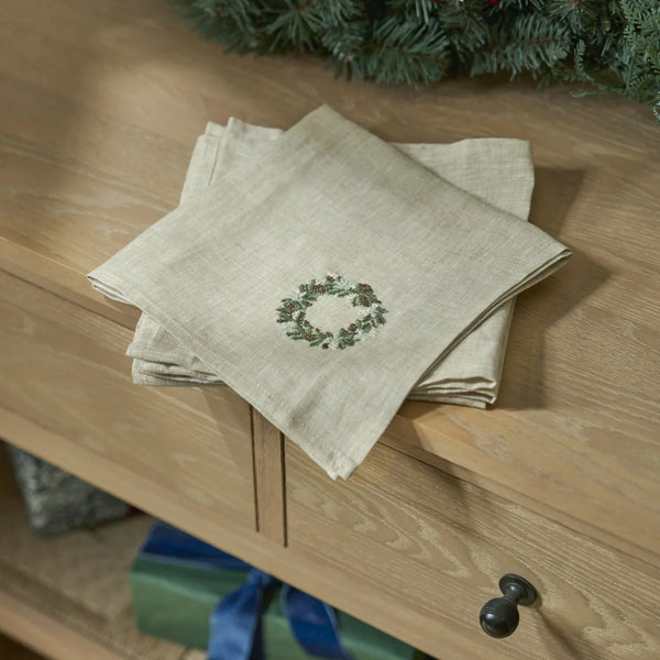 A stack of light beige linen napkins with a green holly wreath embroidery, styled on a wooden drawer in a holiday setting.