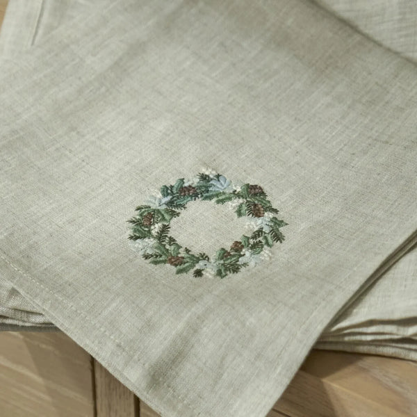 A close-up of a light beige linen napkin featuring an embroidered green and white wreath design, resting on a wooden surface.