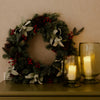 A festive green pine wreath decorated with red berries, silver eucalyptus leaves, and pinecones, displayed on a dark wooden mantel next to two lit amber glass candle holders against a textured beige wall.
