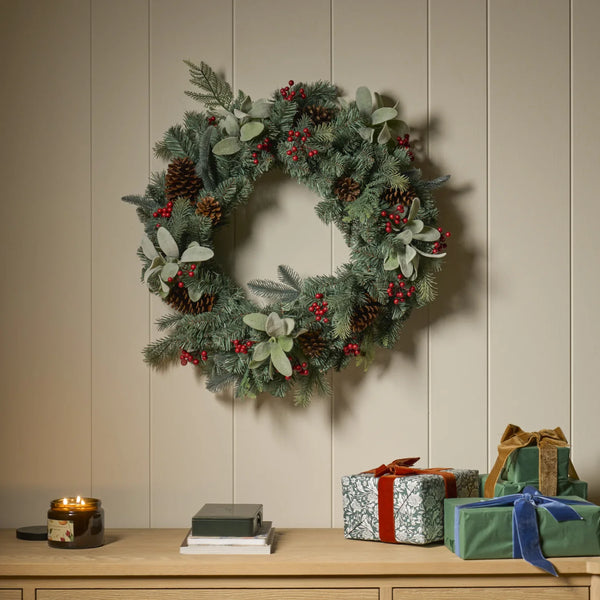 A festive green wreath decorated with pinecones, red berries, and eucalyptus leaves, hanging on a beige paneled wall above a wooden console table with wrapped gifts and a lit candle.