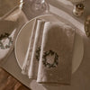 A set of beige linen napkins with embroidered green and white floral wreaths, folded on white plates on a table setting.