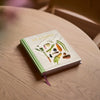 A hardcover book titled "SEASONS" resting on a light wood table. The cover is off-white with illustrations of various vegetables and herbs, and the spine and top edge are light green.
