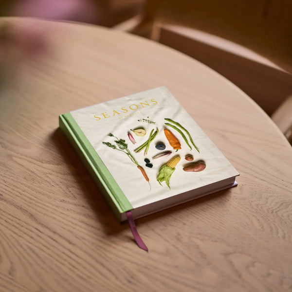 A hardcover book titled "SEASONS" resting on a light wood table. The cover is off-white with illustrations of various vegetables and herbs, and the spine and top edge are light green.