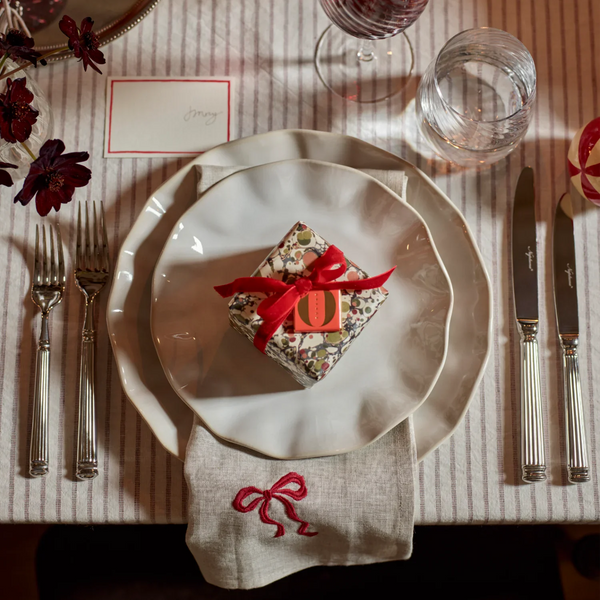 A styled table setting with layered white plates, silver flatware, a striped tablecloth, and a small wrapped gift box tied with a red ribbon in the center. A linen napkin with an embroidered red bow sits below the plates, and glassware and flowers are visible around the setting.