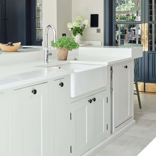 A white farmhouse kitchen sink installed in a white shaker-style island cabinet, shown in a bright modern kitchen with a chrome gooseneck faucet, potted plants, and dark blue paneled doors in the background.